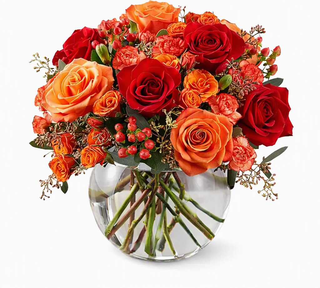 A vibrant autumn floral arrangement featuring red and orange roses, orange carnations, and small red berries in a clear glass vase, set against a plain white background.