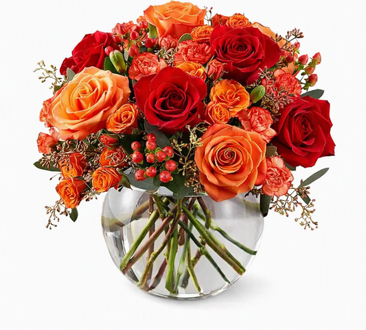 A vibrant autumn floral arrangement featuring red and orange roses, orange carnations, and small red berries in a clear glass vase, set against a plain white background.